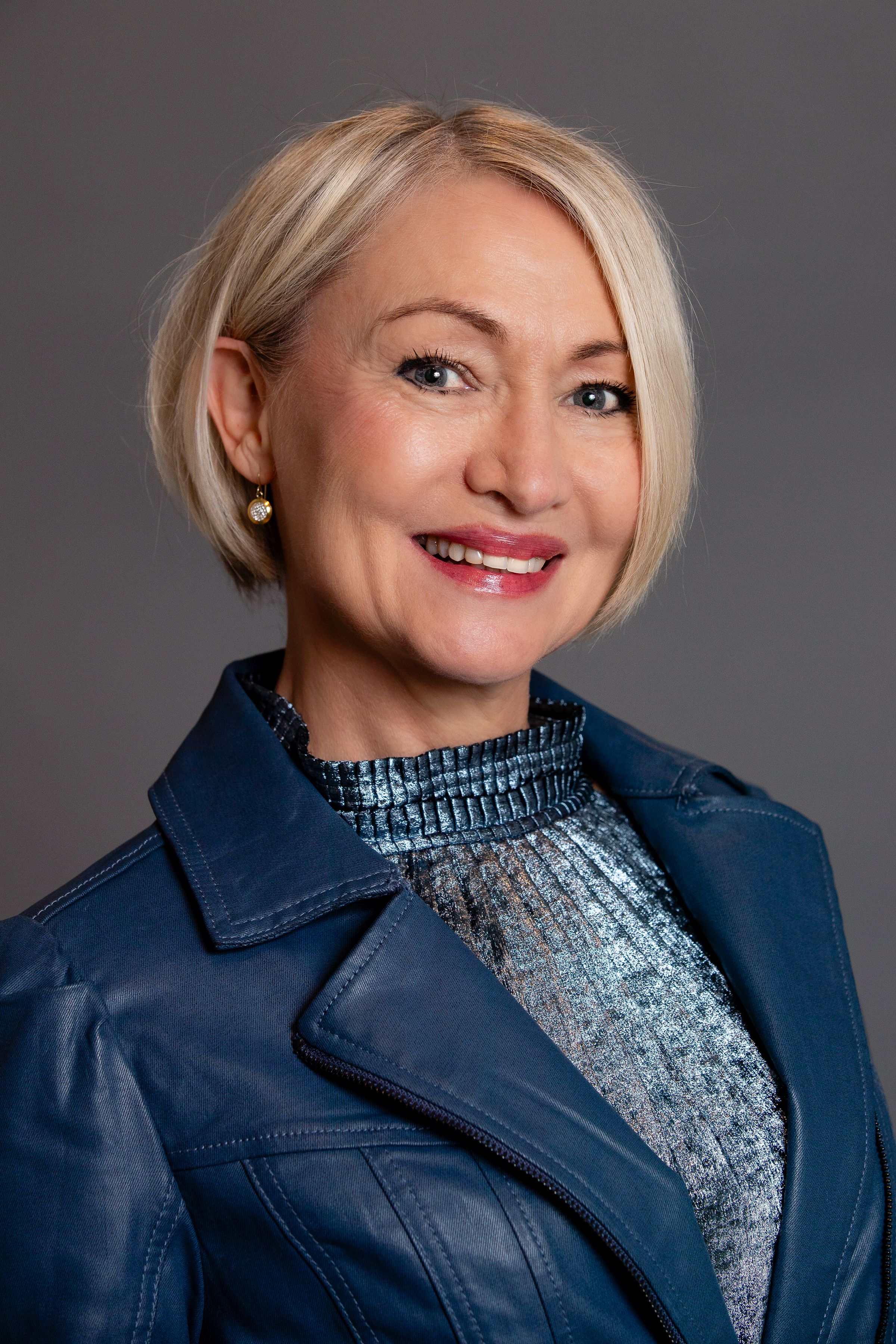 Professional studio portrait showing warm smile wearing navy leather jacket and sparkly silver top against dark background.