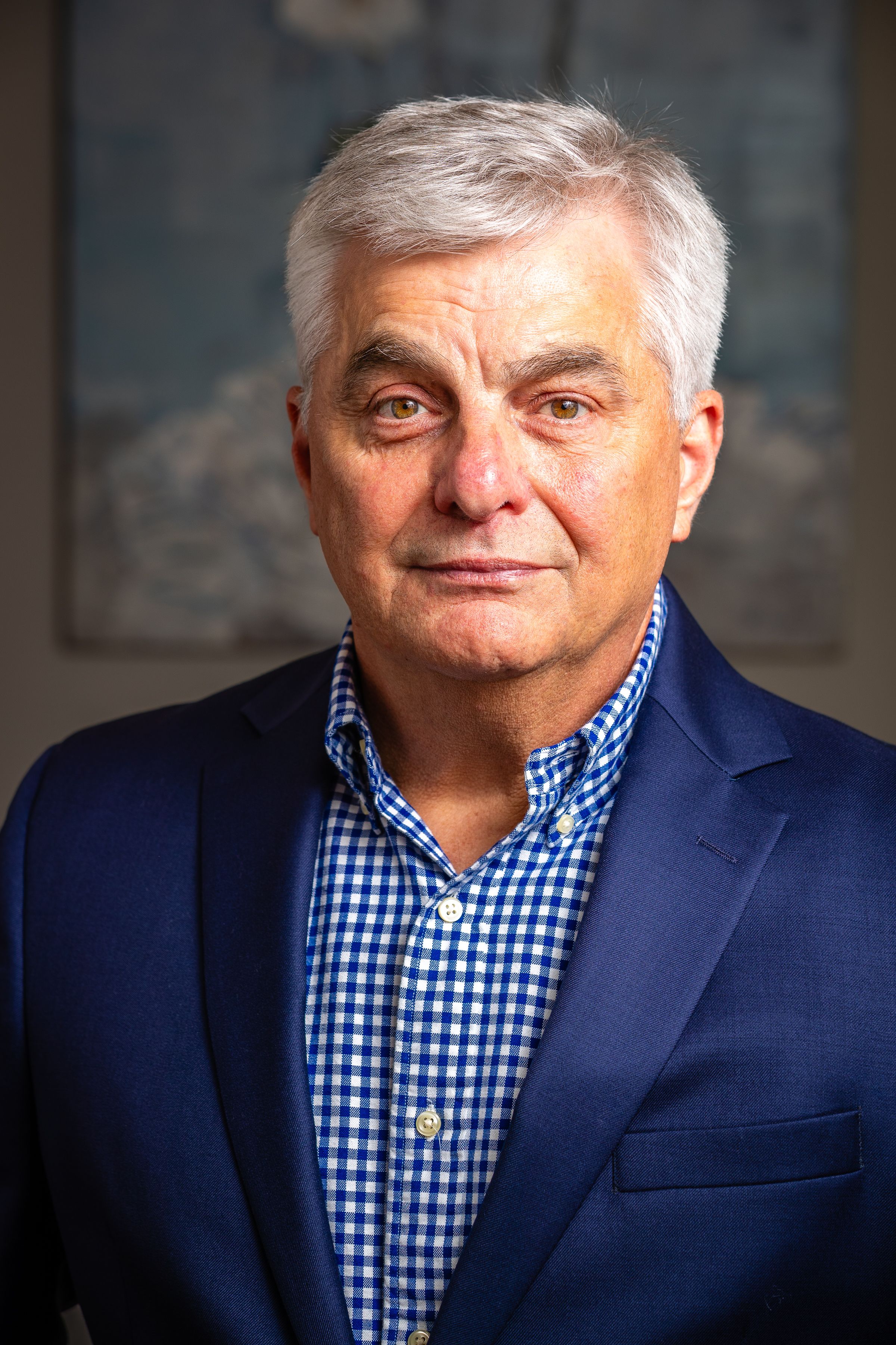 Portrait of a mature executive in a navy blazer and blue gingham check shirt against a blurred background.