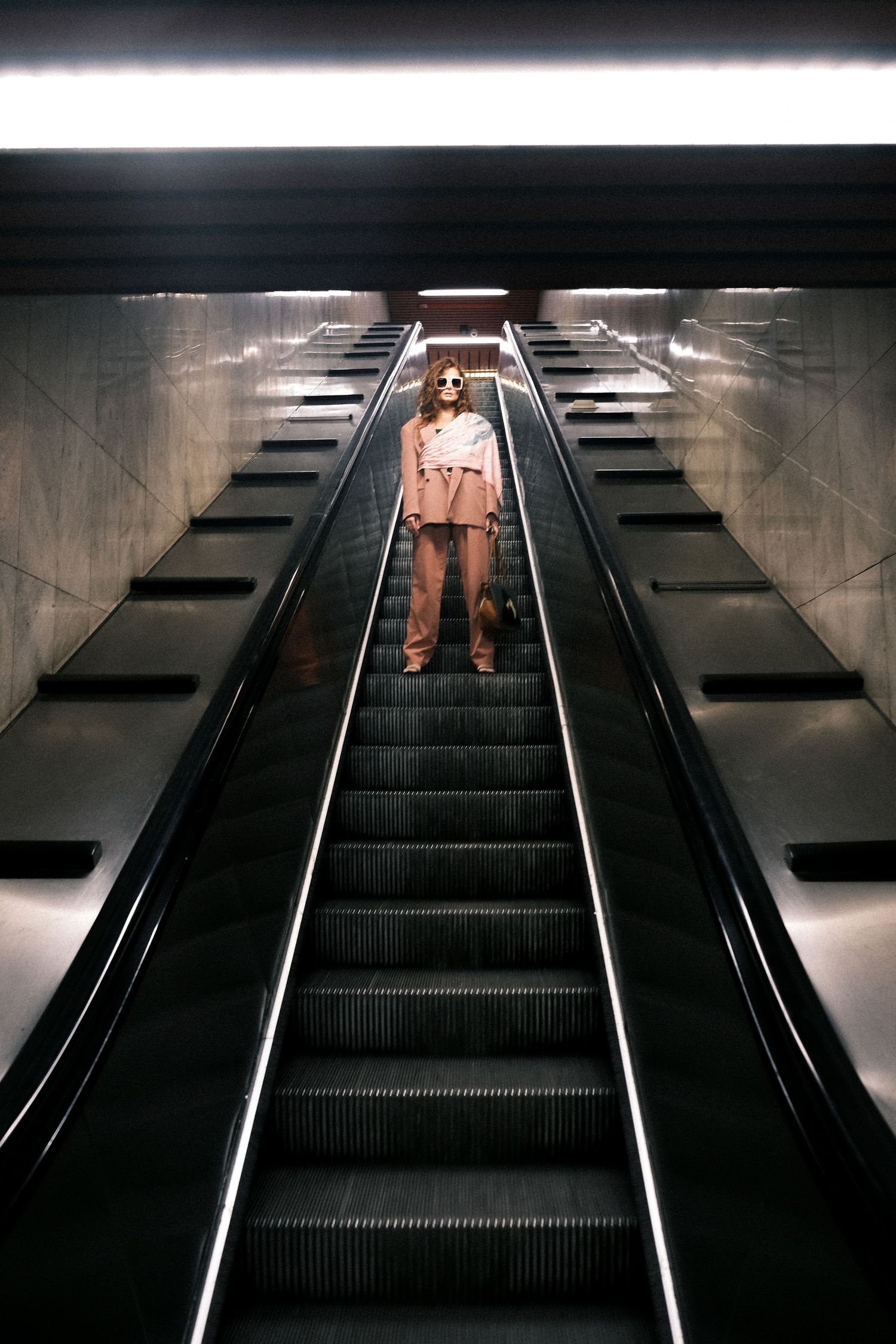 Person in tan outfit standing alone on illuminated escalator in dark subway station.