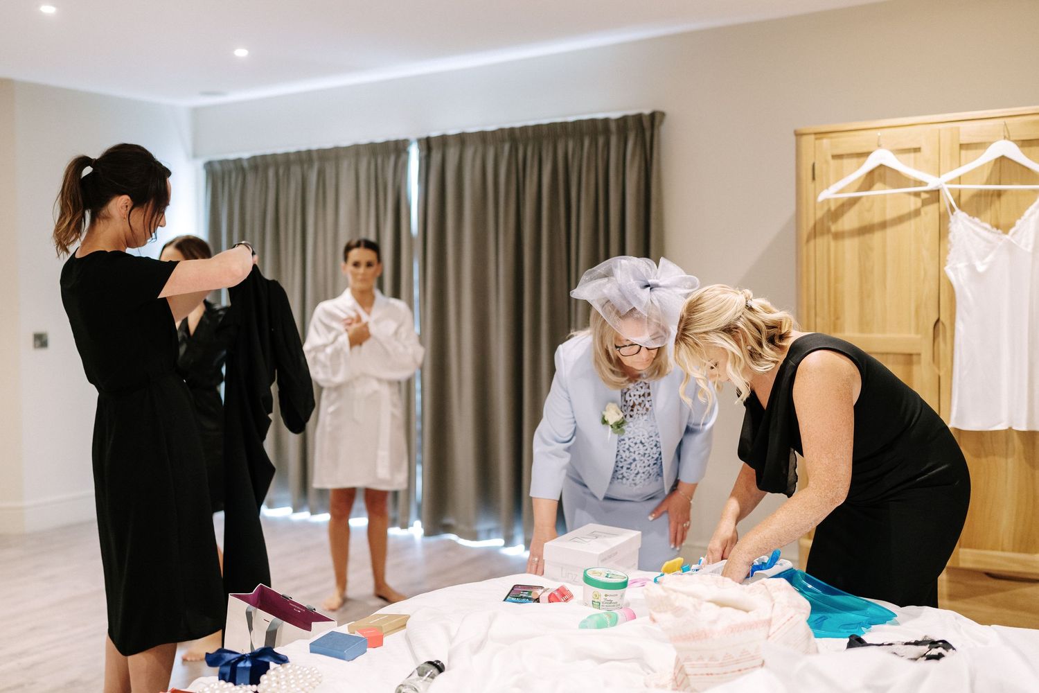 A group of people prepare wedding attire and accessories in a bright hotel suite dressing room.