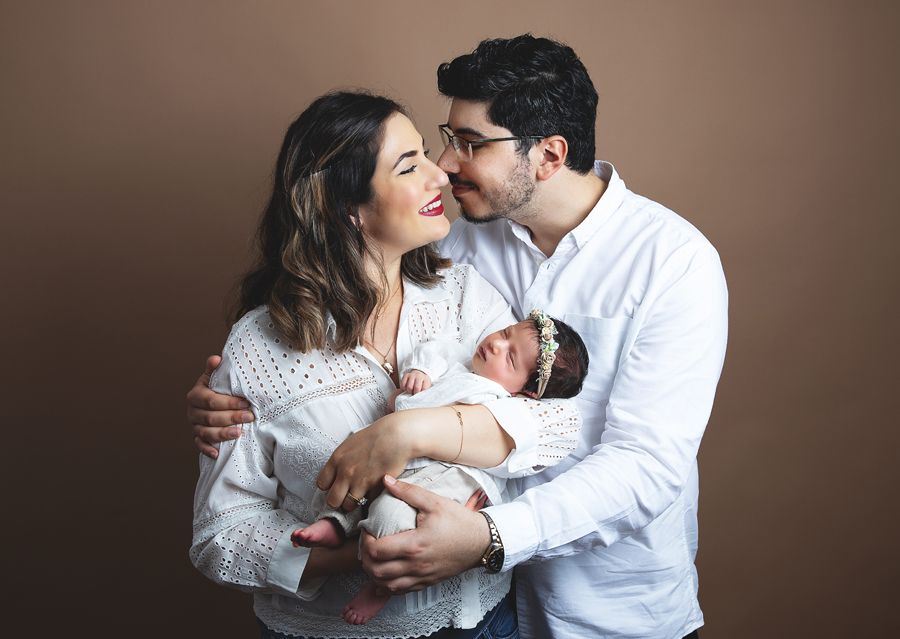 At a newborn photoshoot in London, parents share an intimate moment while holding their newborn baby against a brown background.