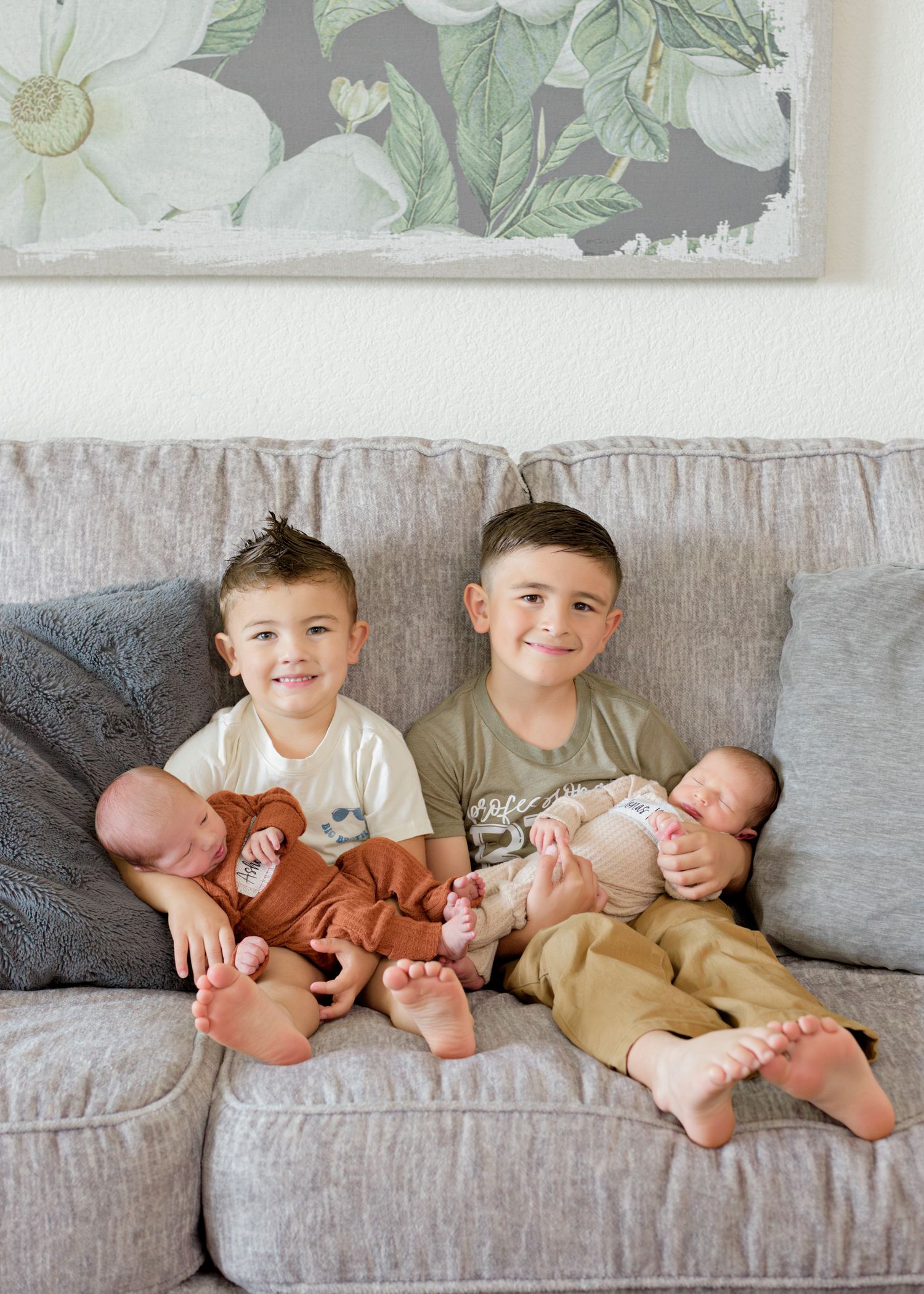 Two young boys on a couch cuddle with infant babies while sitting beneath floral wall art.
