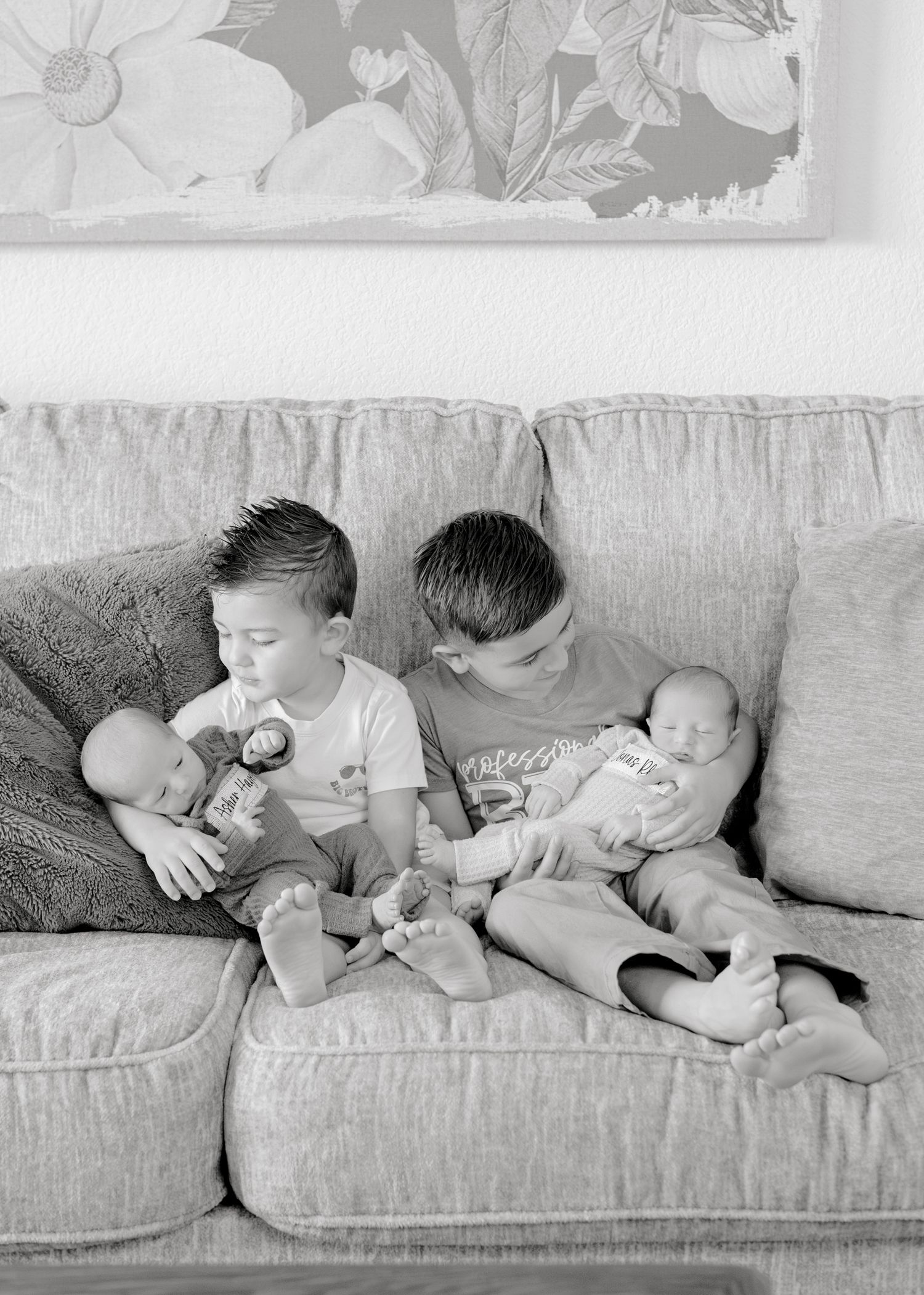 Two siblings sitting on a gray couch hold and feed their infant siblings in a tender black and white moment.