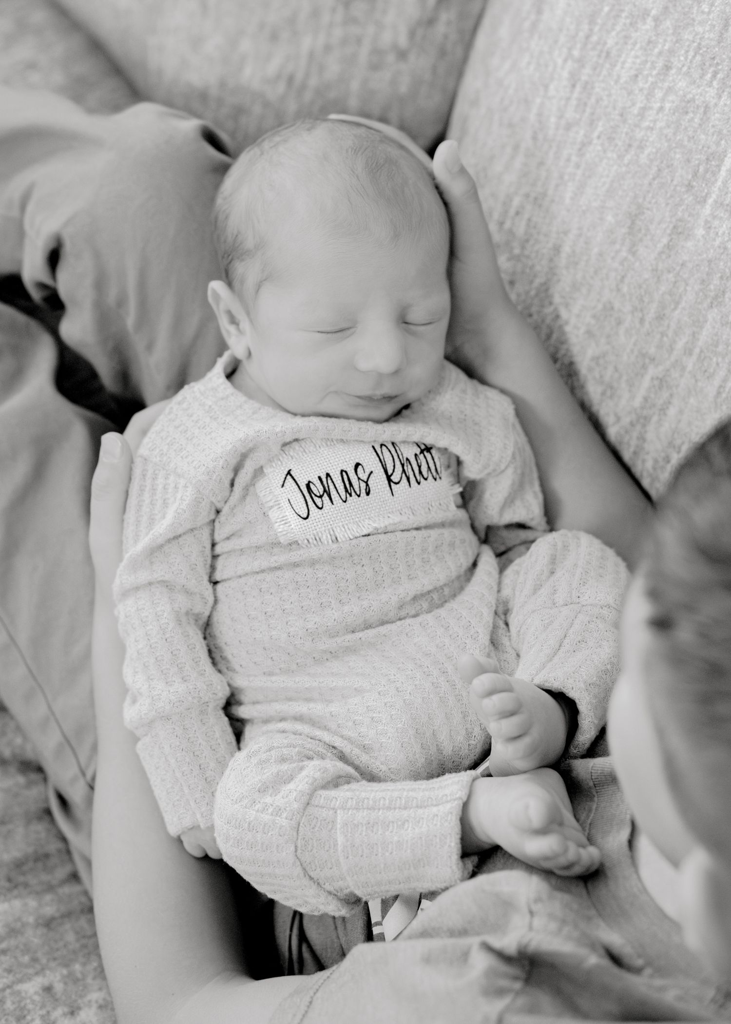 A sleeping newborn wearing a gray onesie lying on a soft cushion in black and white.