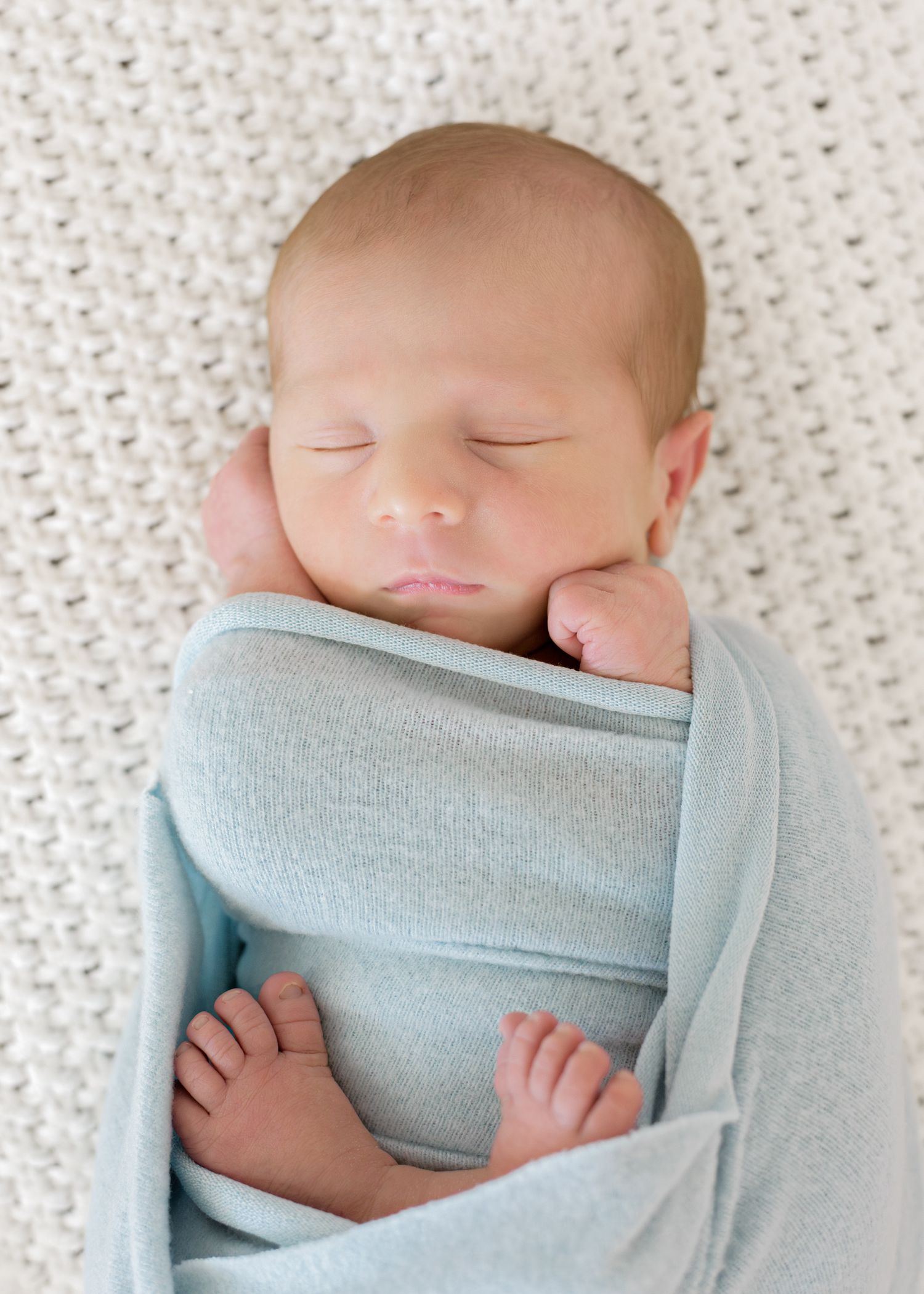 Sleeping newborn baby wrapped in light blue swaddle blanket on textured white background.