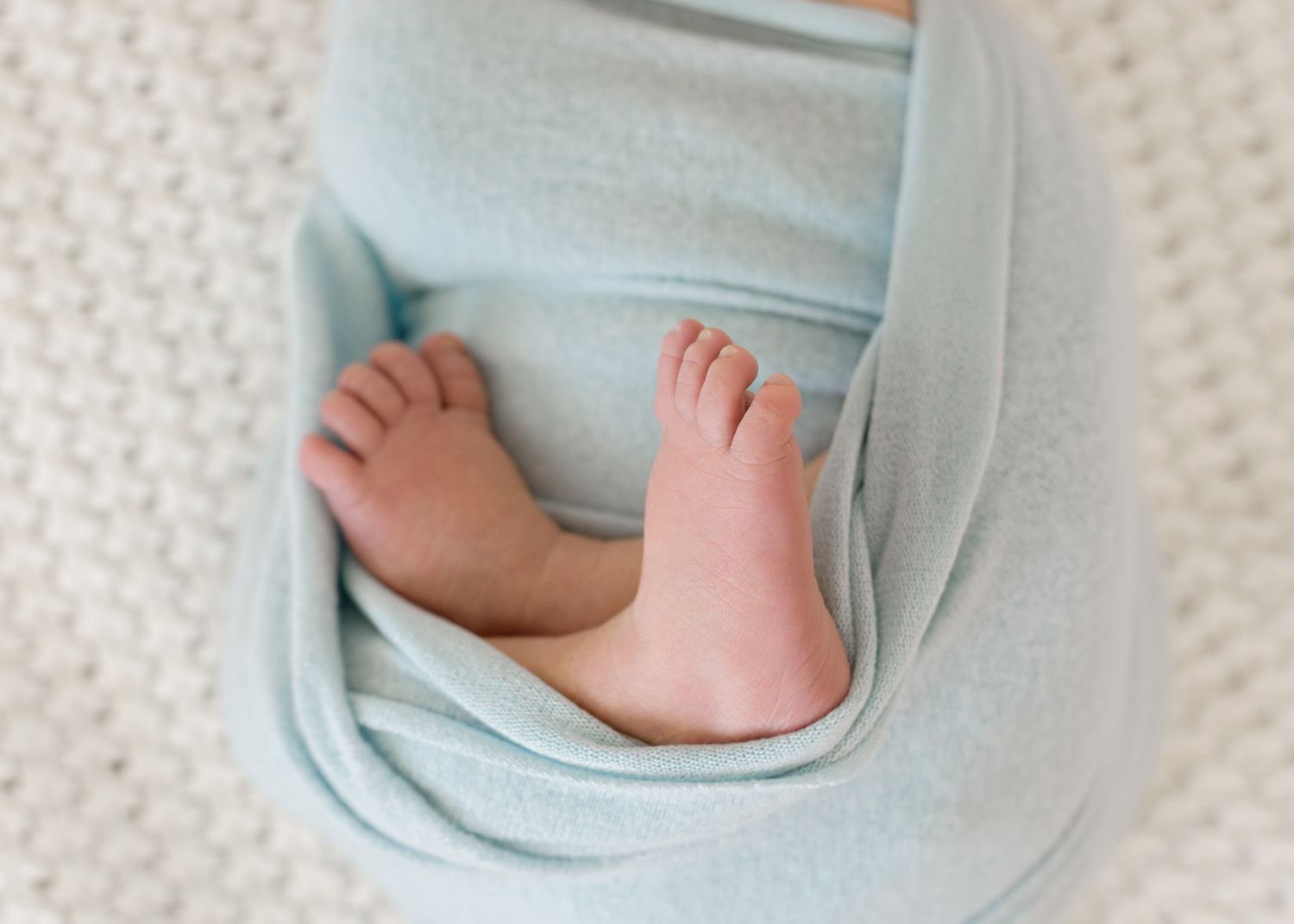 Tiny pink baby feet peeking out from under a soft light blue blanket against white textured fabric.