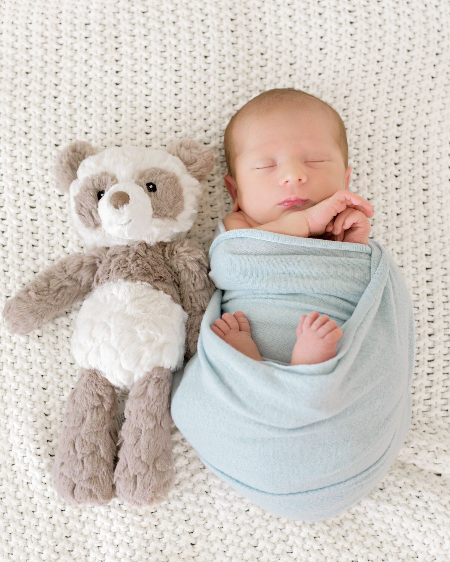 Sleeping baby swaddled in light blue blanket lying on textured white bedding next to plush teddy bears.