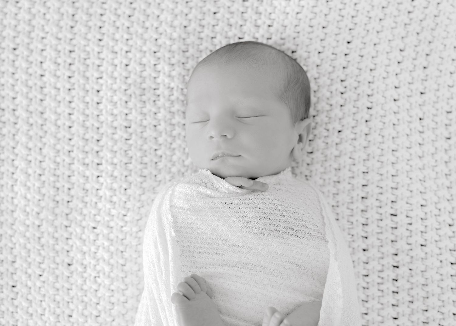 An infant rests peacefully on a grey and white knitted blanket with patterned stitching.