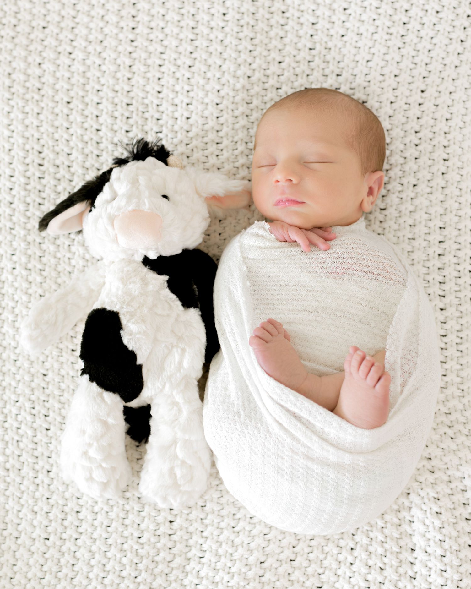 Sleeping infant in white swaddle lying next to black and white plush stuffed cow toy on textured blanket.