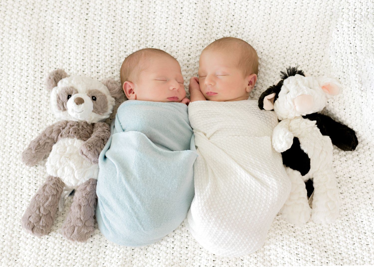 Two sleeping babies lie side by side on white bedding with soft gray teddy bears.