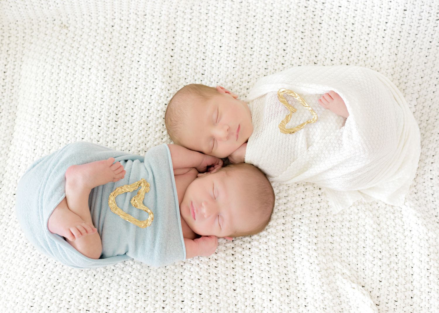 Two sleeping newborns in white and blue onesies with gold designs lie together on a knitted blanket.