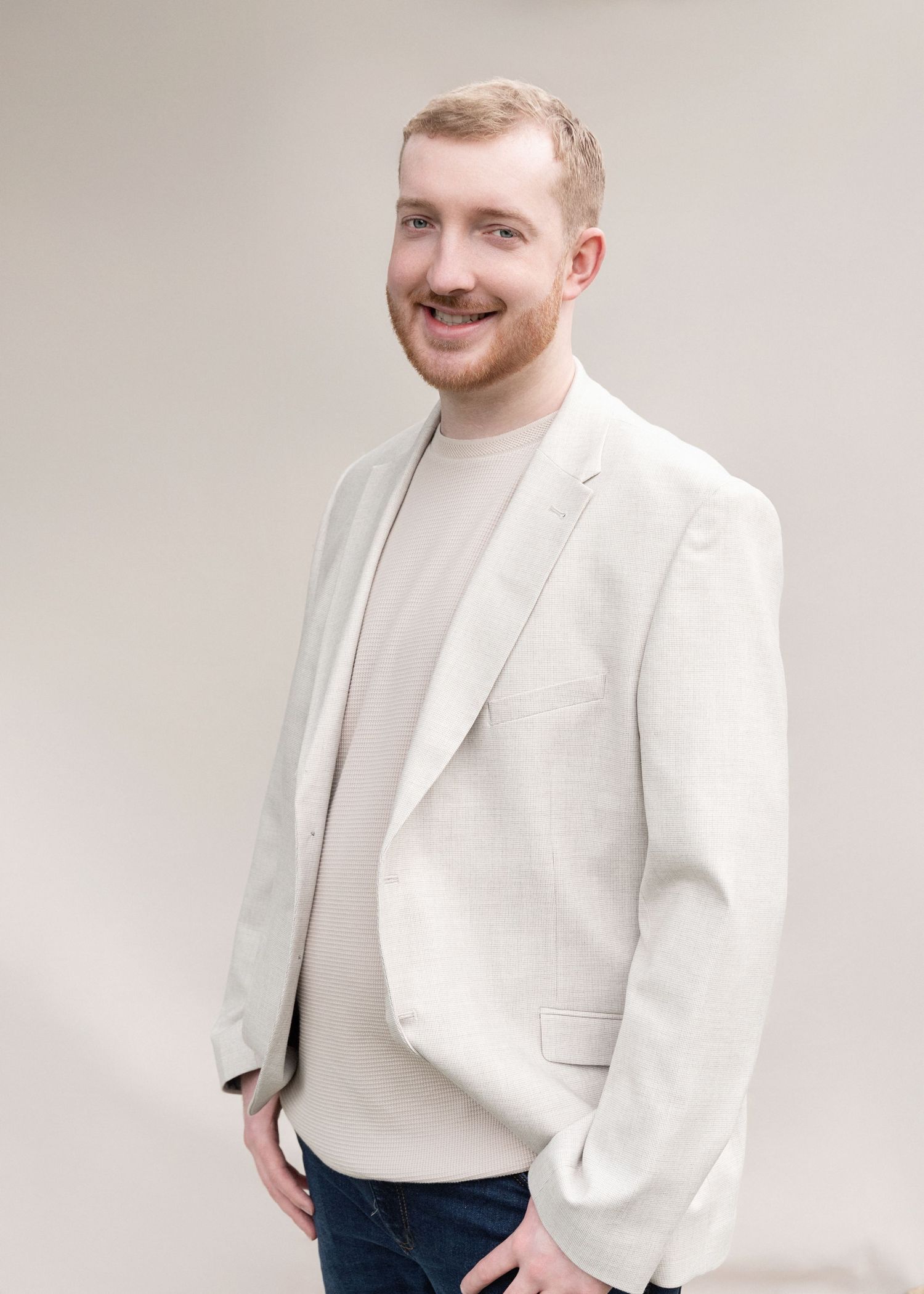 Person wearing a white blazer and black pants poses against a light background while smiling and gesturing.
