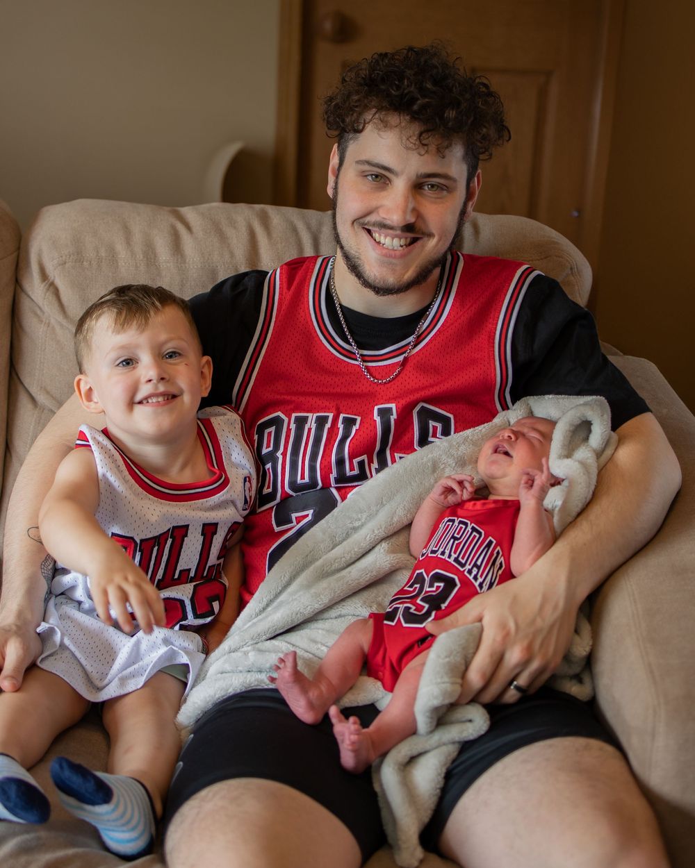 Three people wearing matching red Chicago Bulls basketball jerseys sit together on a beige couch.