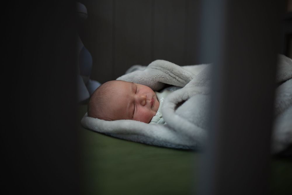 A peaceful sleeping baby nestled in soft blankets viewed through crib bars in dim lighting.