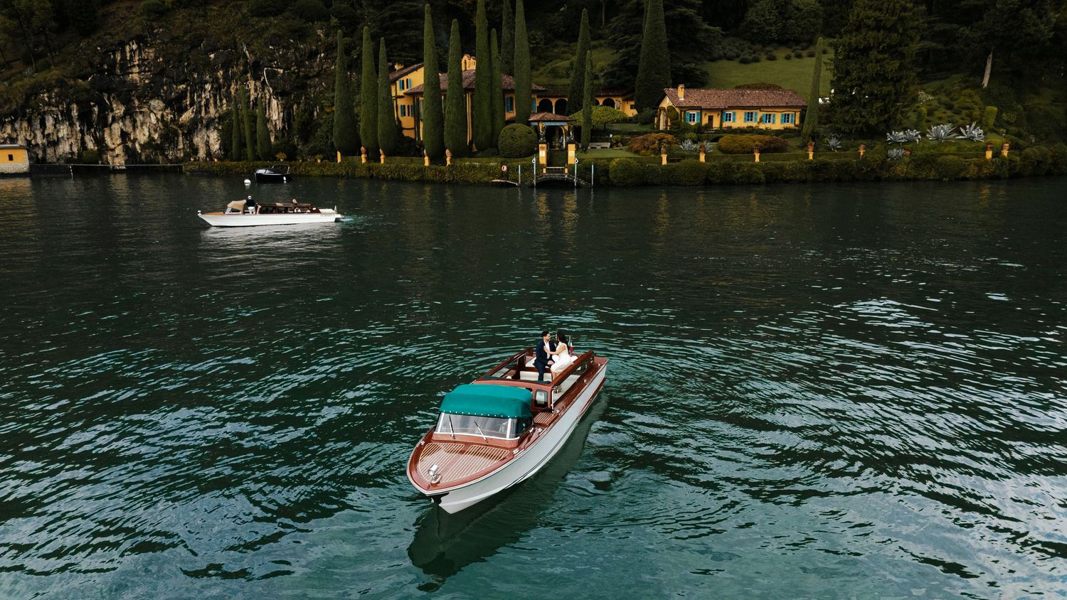 A luxurious wooden boat cruises across the turquoise waters of Lake Como with mountains and Italian villas in the background.