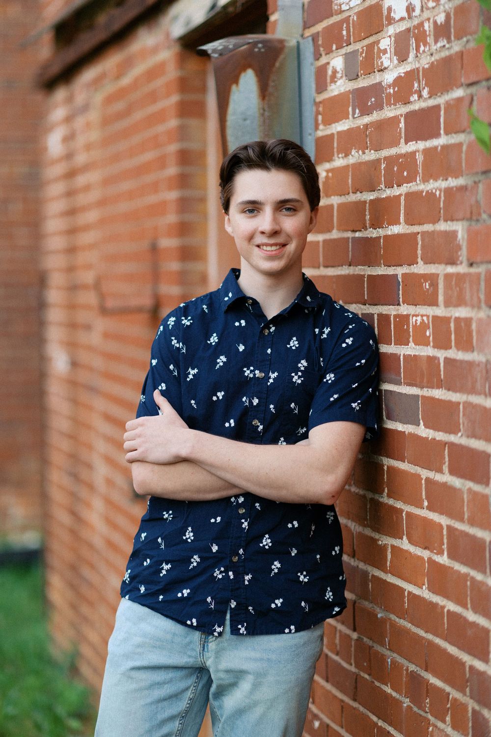 Someone stands confidently against a brick wall wearing a navy patterned short-sleeve shirt and light jeans.