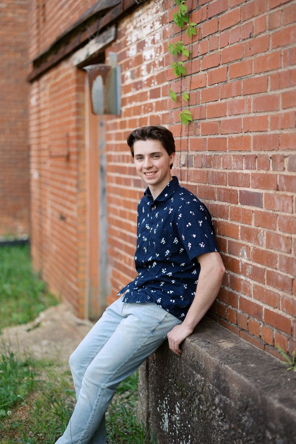 Someone in a navy patterned shirt and light jeans leans casually against a brick wall on a sunny day.