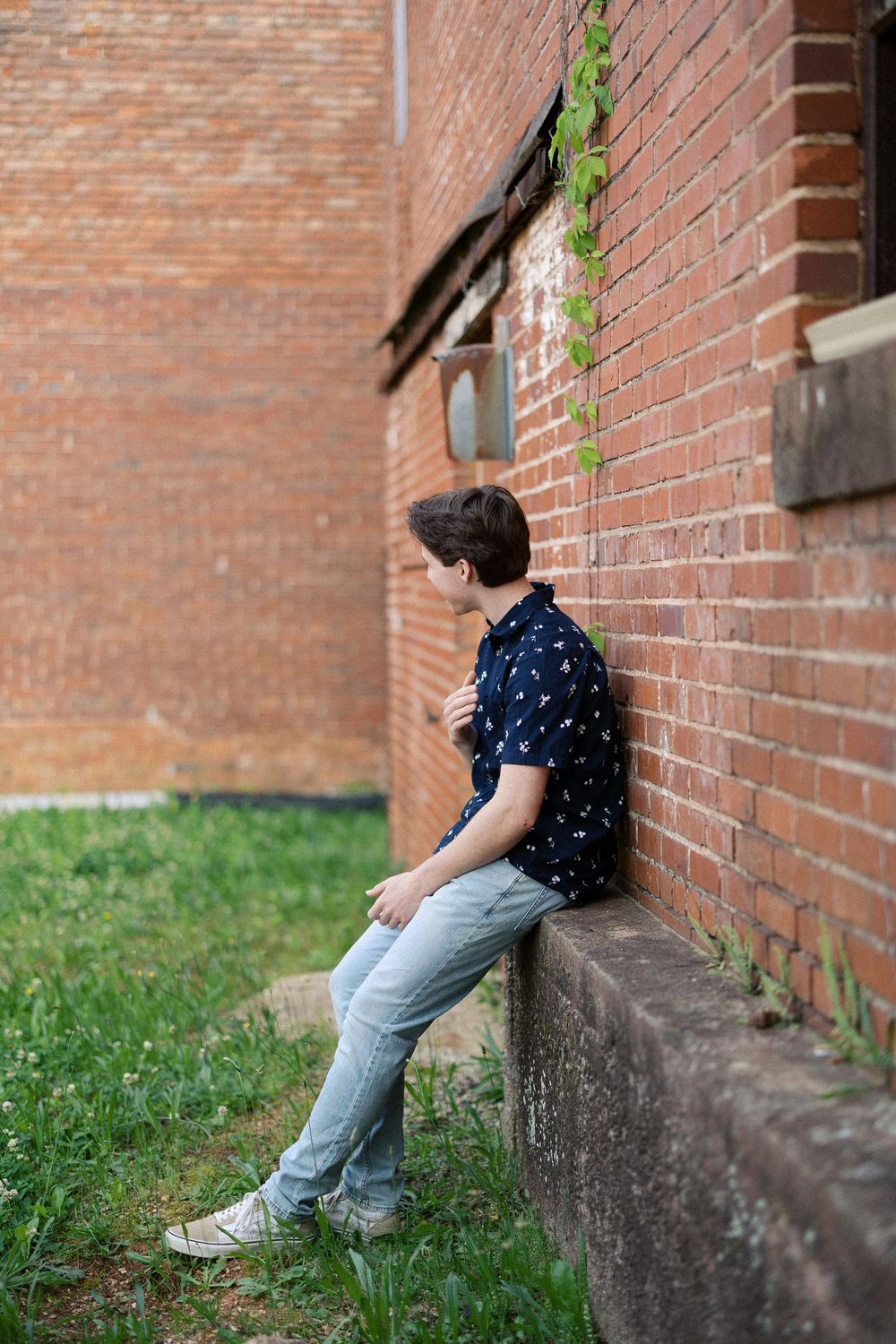 Someone in a navy patterned shirt and jeans leans against a brick wall outside a building with ivy growing above.
