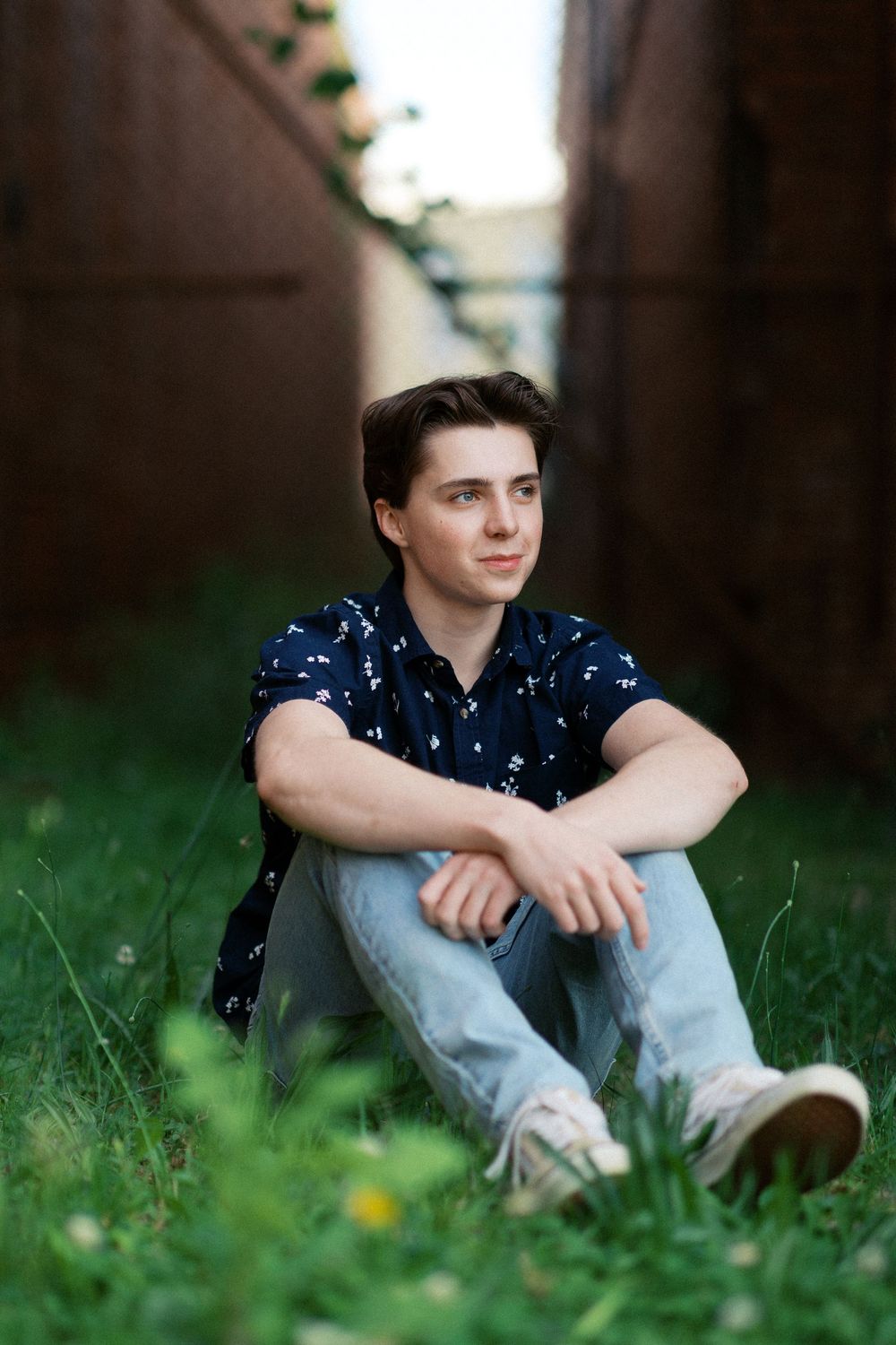 A young person in a navy polka dot shirt and jeans sits thoughtfully in green grass with dandelions.