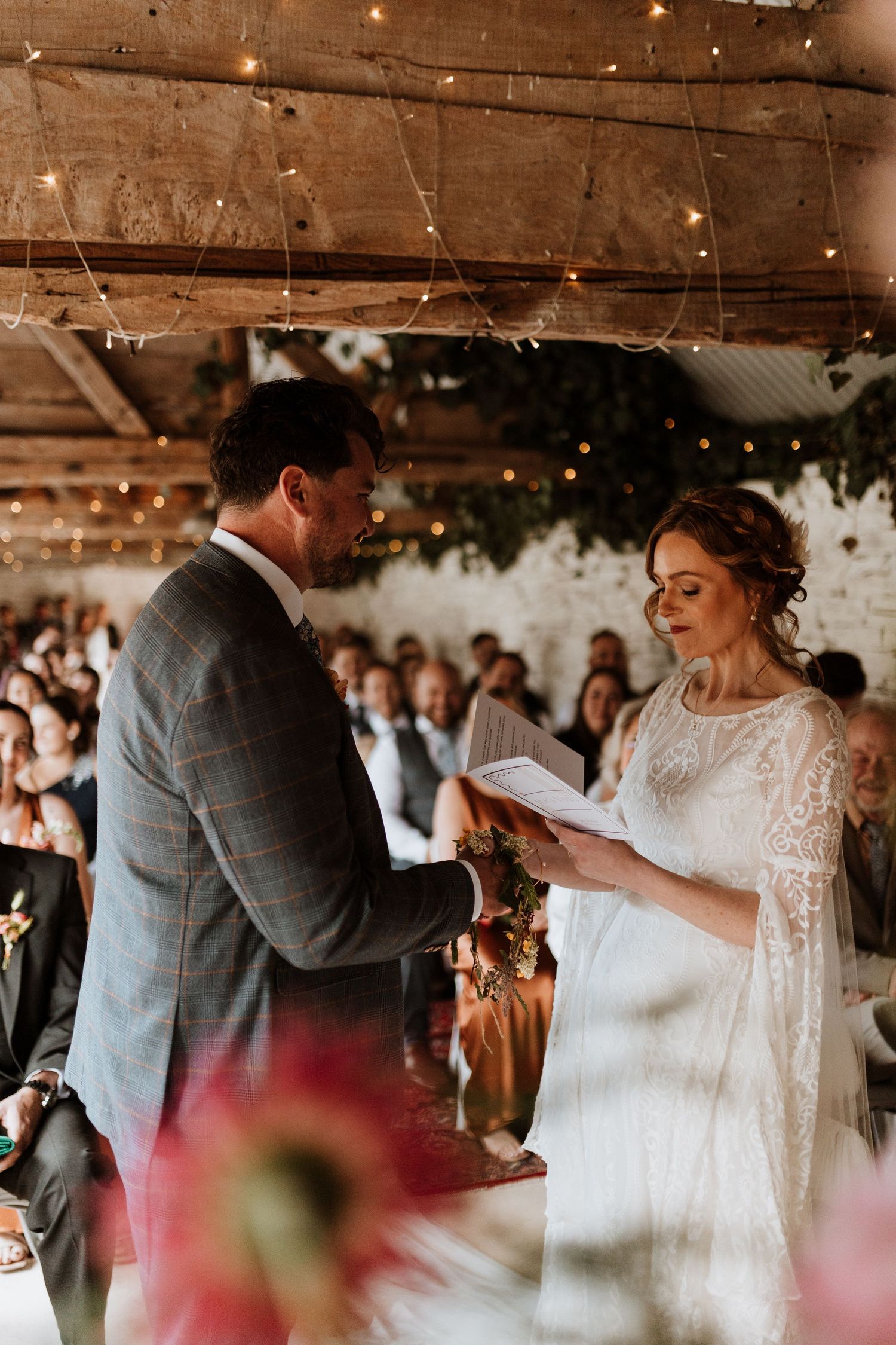 A tender moment shared by newlyweds exchanging vows at their rustic barn wedding reception with string lights overhead.