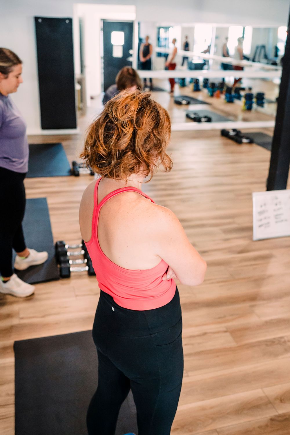 Person in pink tank top and black leggings stands in a bright fitness studio with mirrors and wood floors.
