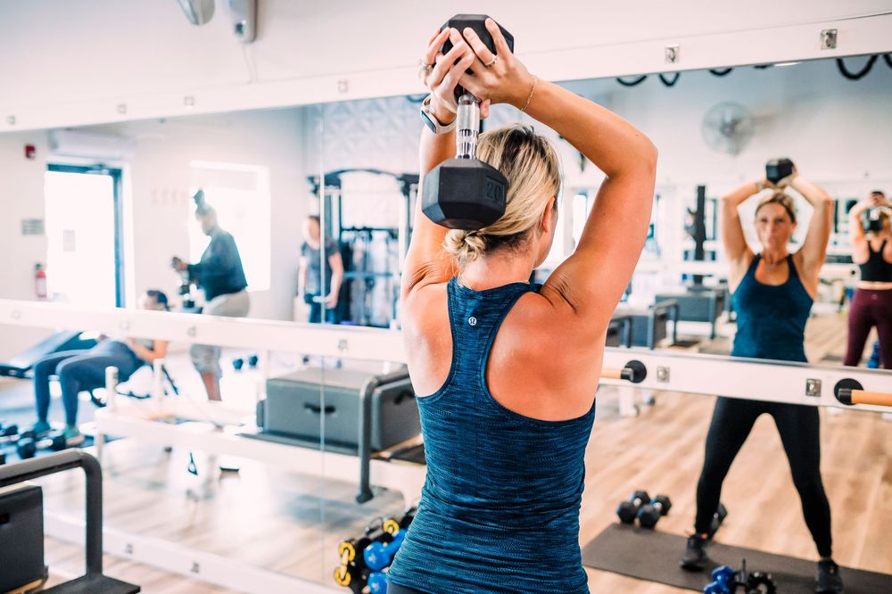Person lifting weights overhead during group fitness class in mirrored gym.