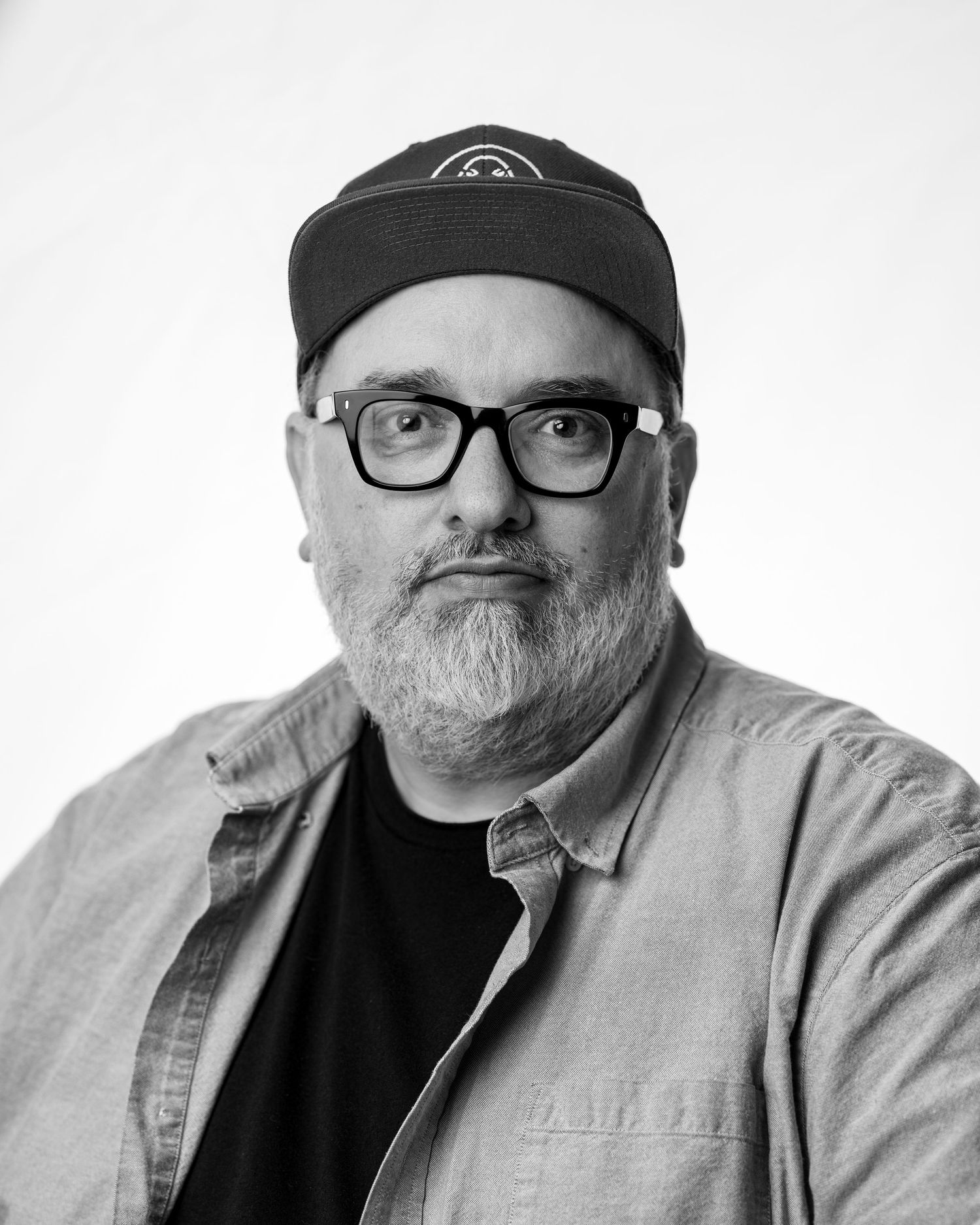Black and white portrait of someone wearing glasses, a baseball cap, and casual attire looking at the camera.