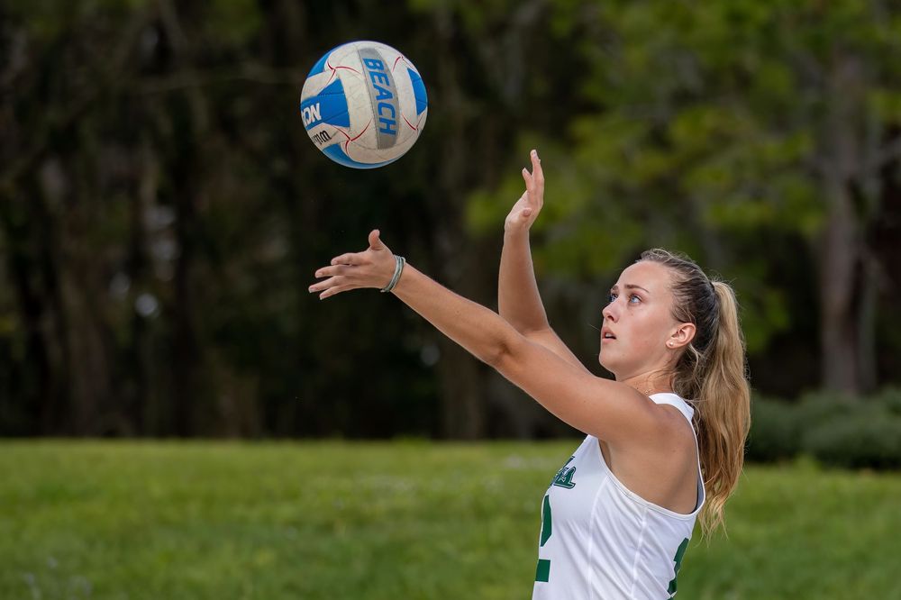 Volleyball player serves ball during outdoor match in sunny park setting.