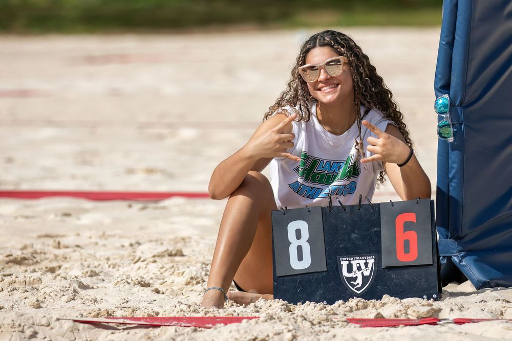 Beach volleyball players on sandy court with scoreboard visible in background.
