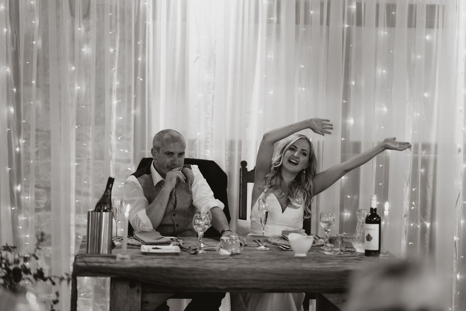 Black and white photo of two people at a dinner table in a celebratory mood, with wine bottles and curtains in background.