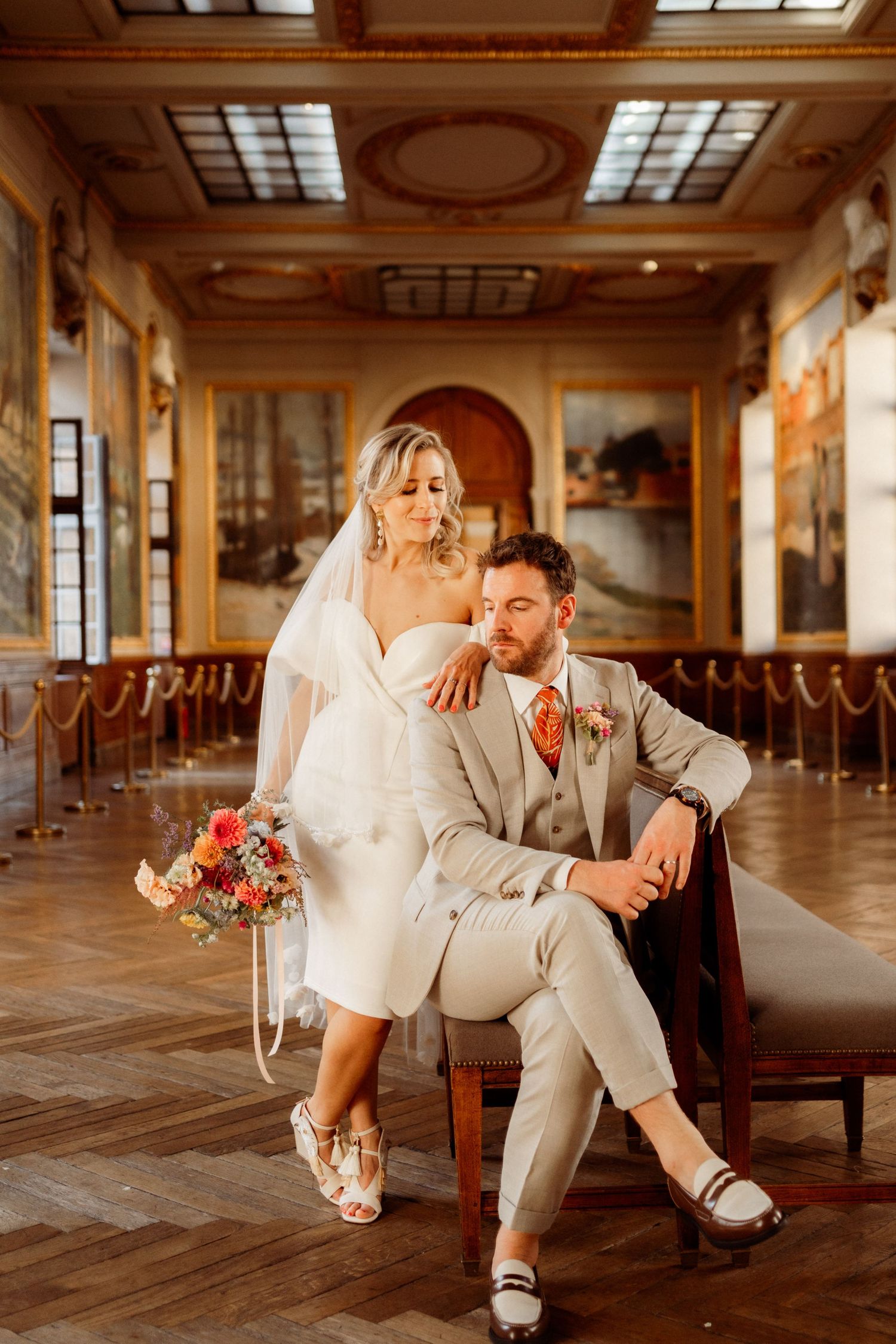 Bride and groom pose elegantly in an ornate hall, she holds a colorful bouquet while he sits in a vintage chair.