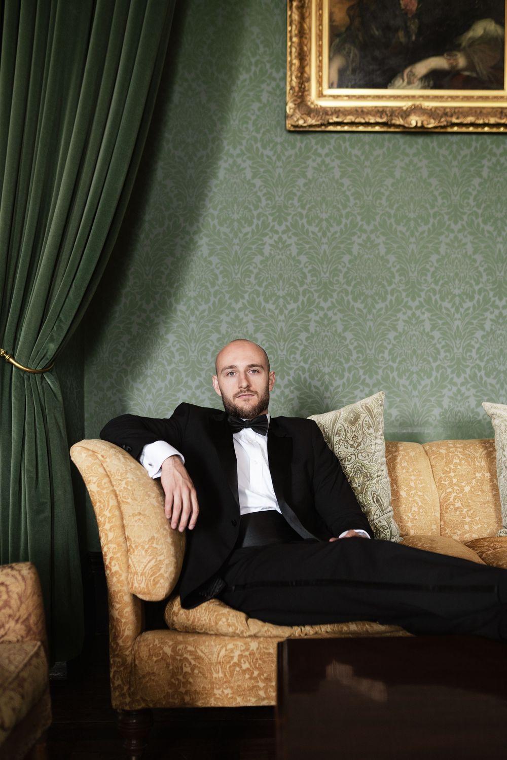 A person in a black suit reclines dramatically across an ornate gold-patterned sofa against a green wall with vintage decor.