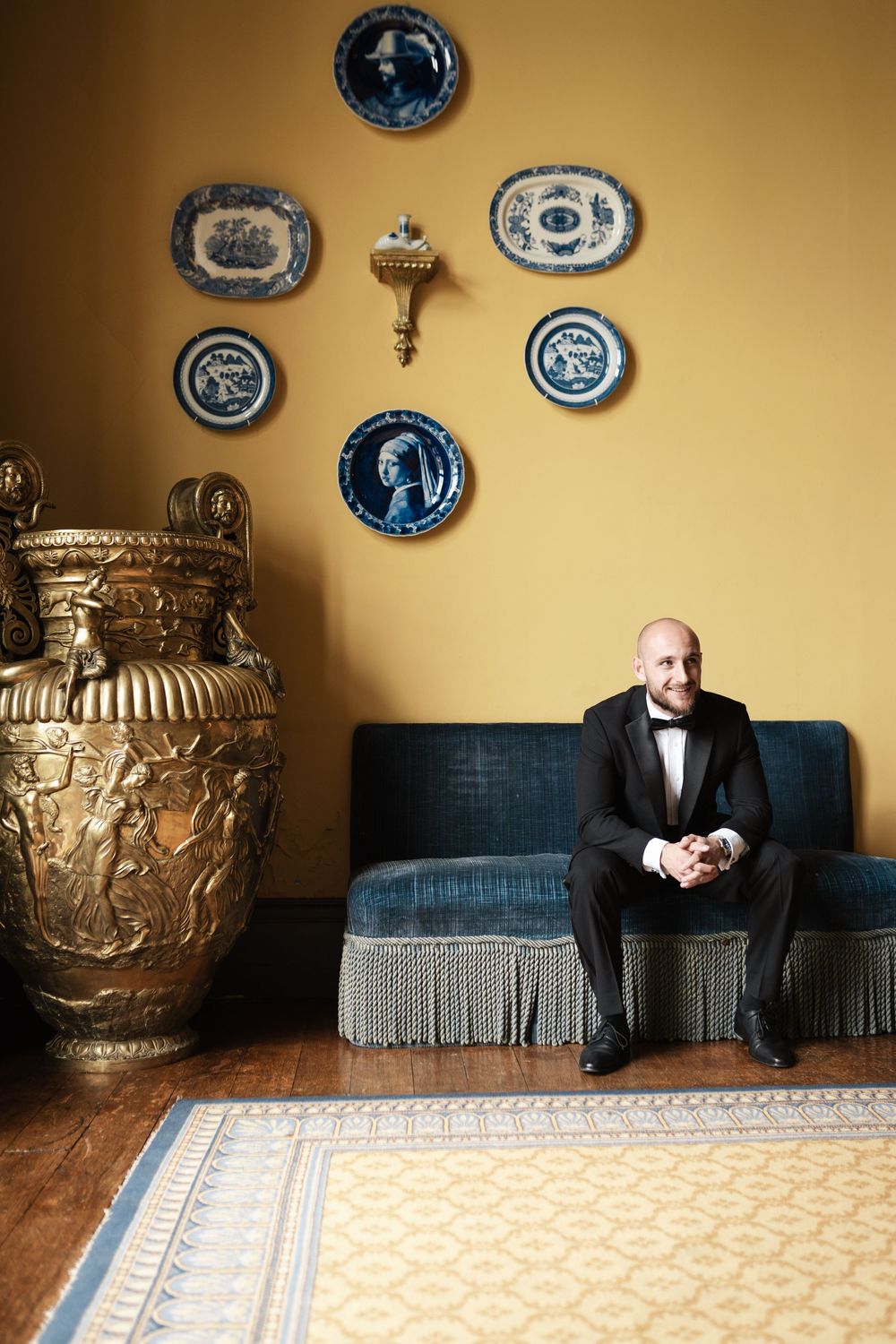 Elegant room interior with decorative plates on yellow wall, large ornate vase, and person seated on blue fringed sofa.