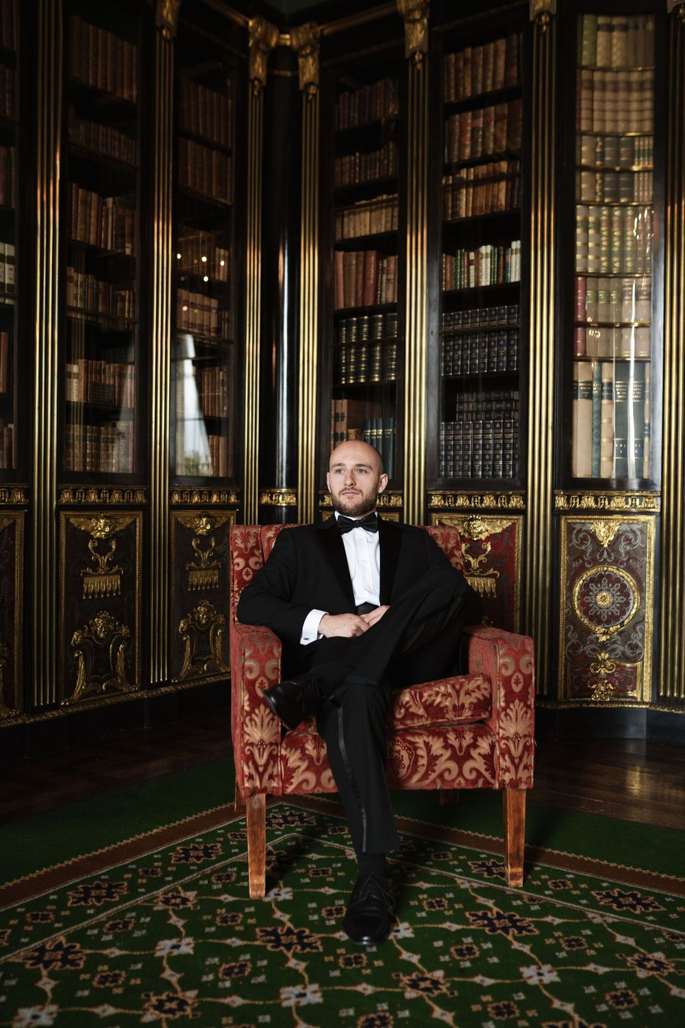 A grand library interior with ornate bookshelves, red velvet chairs, and a green carpet in an opulent historic setting.