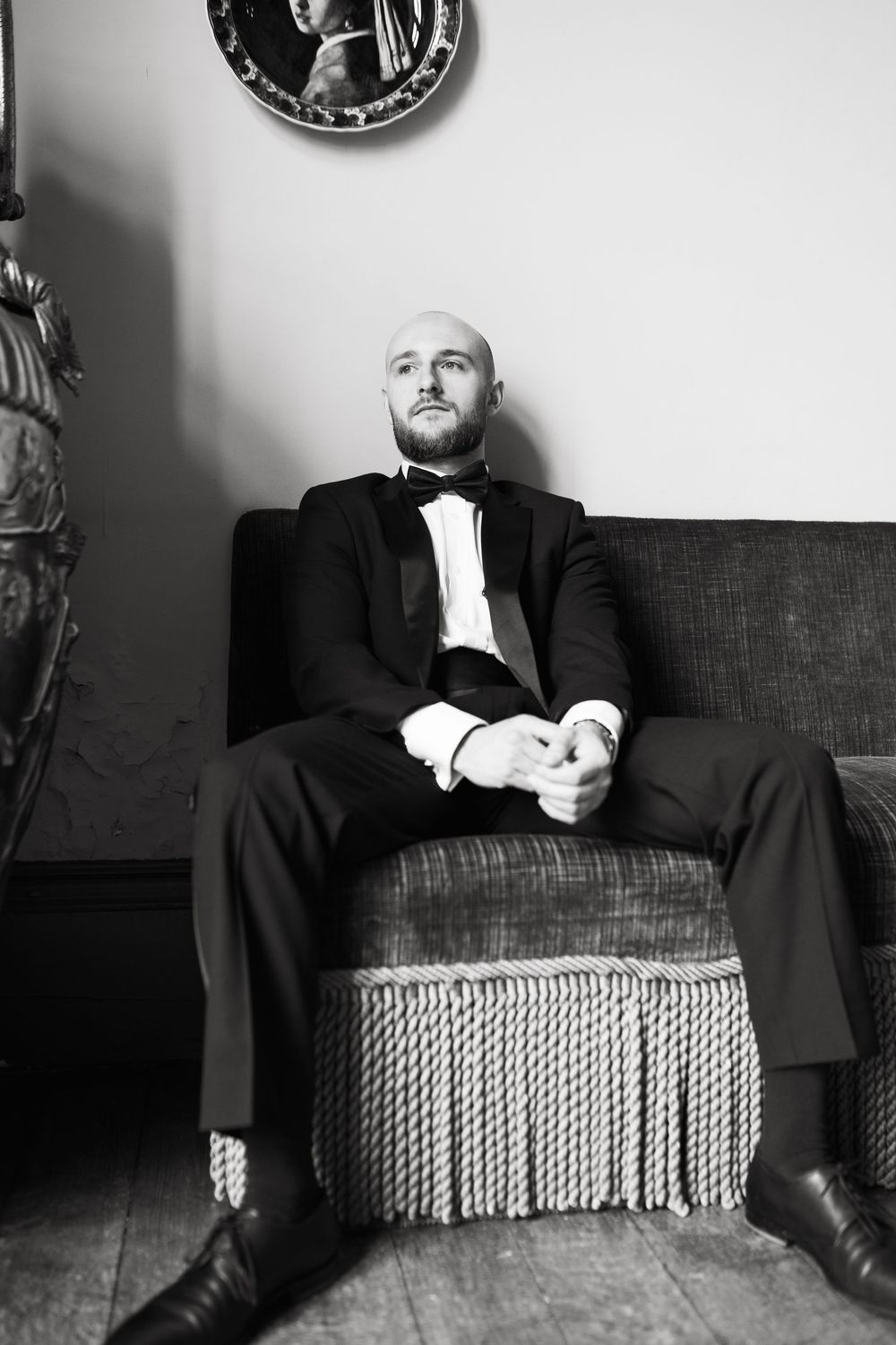 Black and white portrait of someone in formal attire reclining on a vintage fringed armchair in an elegant interior.