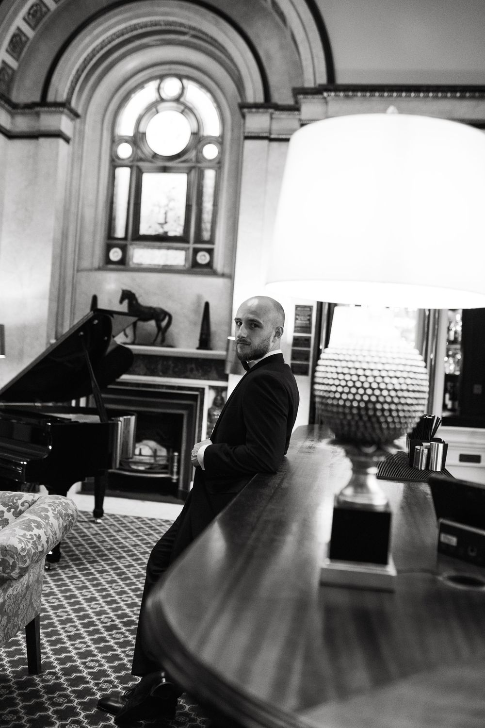 Someone sits alone in a large ornate room with patterned carpet and arched windows in black and white.