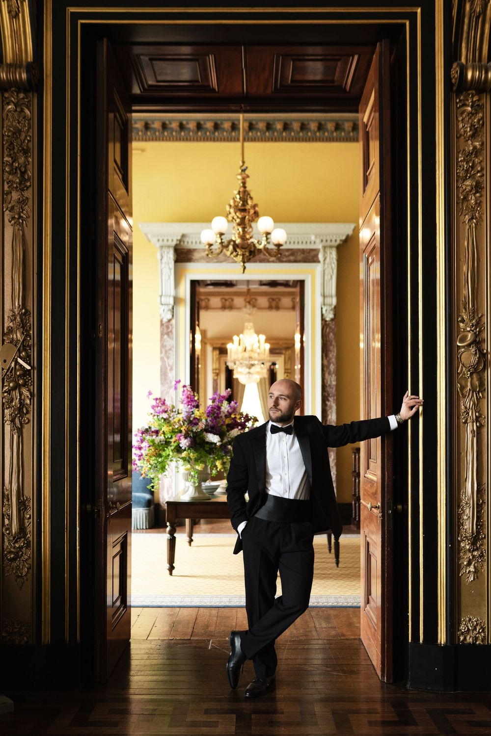 An elegant room in a historic mansion features ornate doorways, chandeliers and floral arrangements in warm golden light.