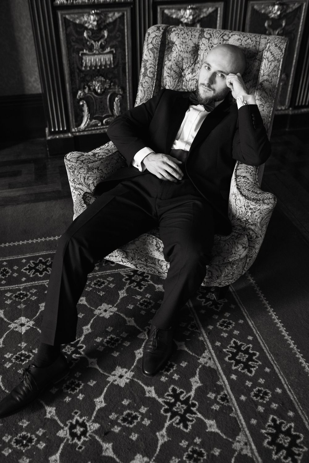 A person wearing a tuxedo reclines elegantly in an ornate armchair on a patterned carpet in black and white.