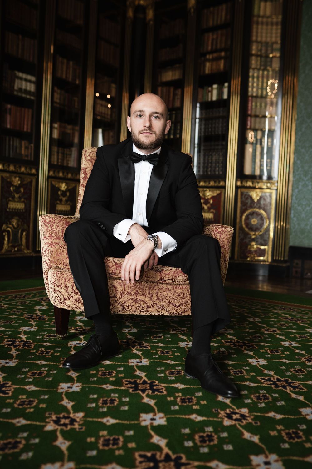 A person in a black tuxedo and bowtie sits elegantly on an ornate chair in a luxurious library with green carpeting.