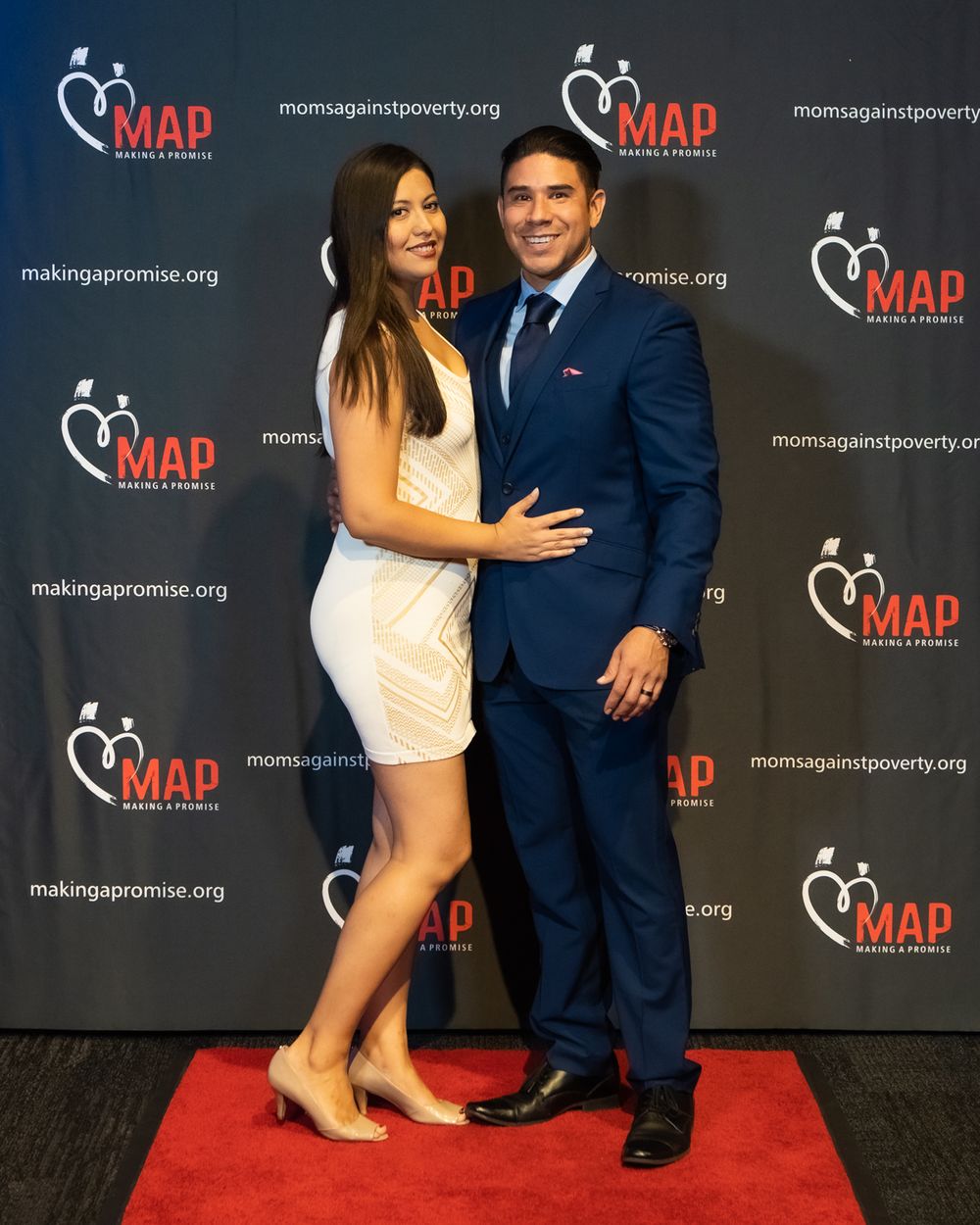 A couple poses together on a red carpet in front of a MAP charity event backdrop, wearing formal attire.