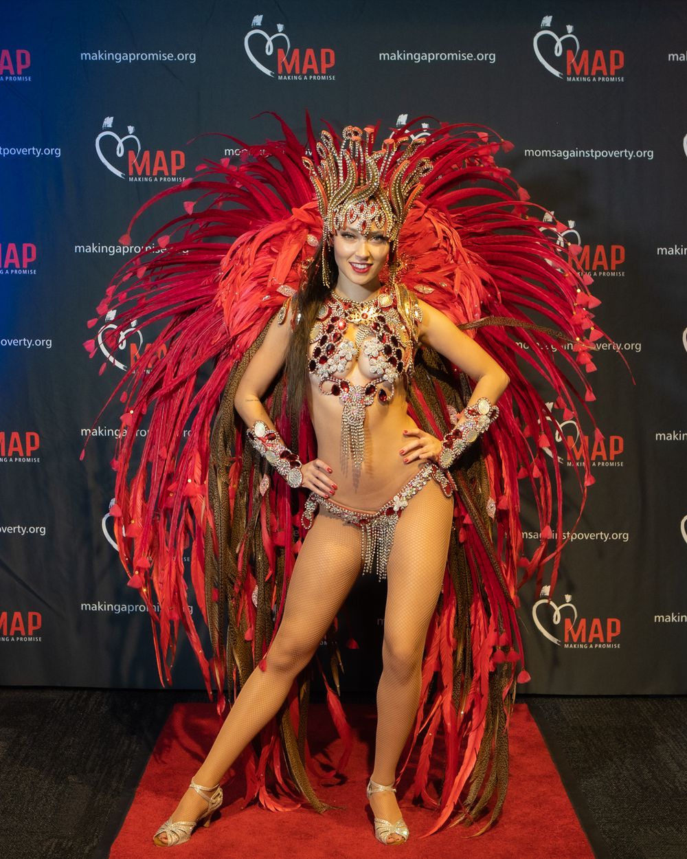 Dancer in bejeweled costume with dramatic red feathered wings poses on red carpet at event.