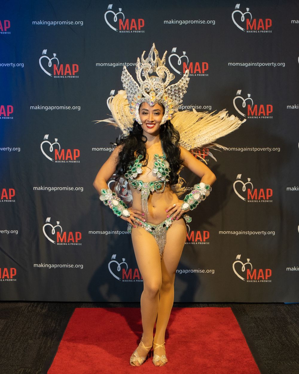 A performer wearing an elaborate gold and silver feathered headdress and sequined costume poses on a red carpet.