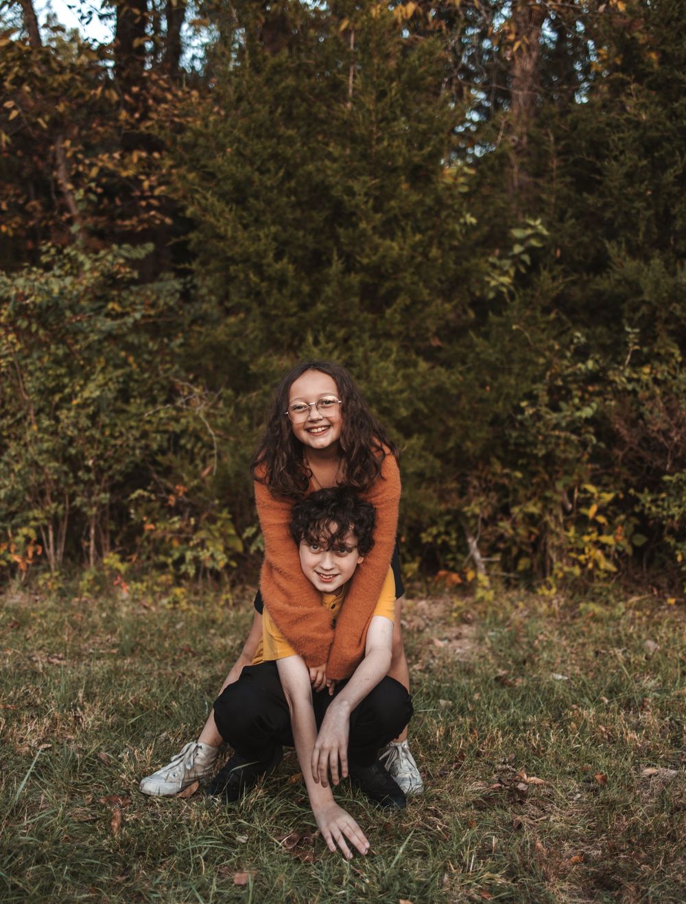 A figure in an orange sweater sits in a relaxed pose on autumn grass with trees in the background.