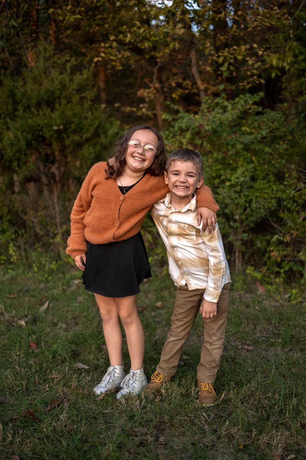 Two people in outdoor casual attire lean towards each other while standing in a grassy area with trees in background.