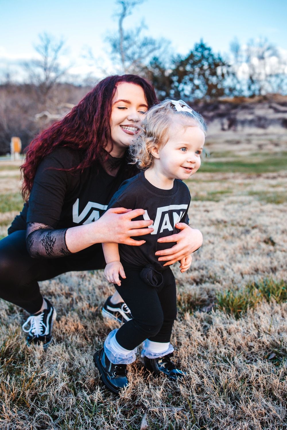 Two people wearing matching black outfits smile and embrace while playing outdoors in a grassy field at dusk.