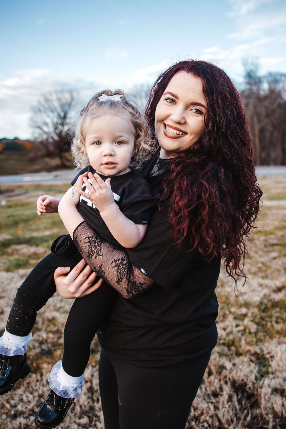 Woman with curly dark hair holding child wearing black outfit in outdoor rustic setting during autumn.