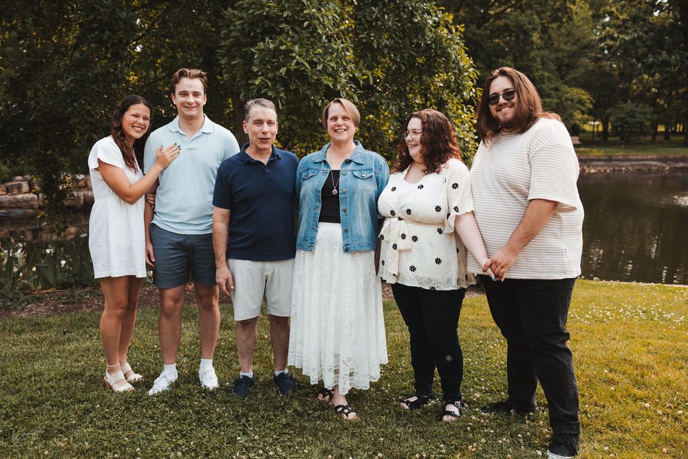 Group gathering on a grassy lawn by a pond on a sunny day wearing casual attire.