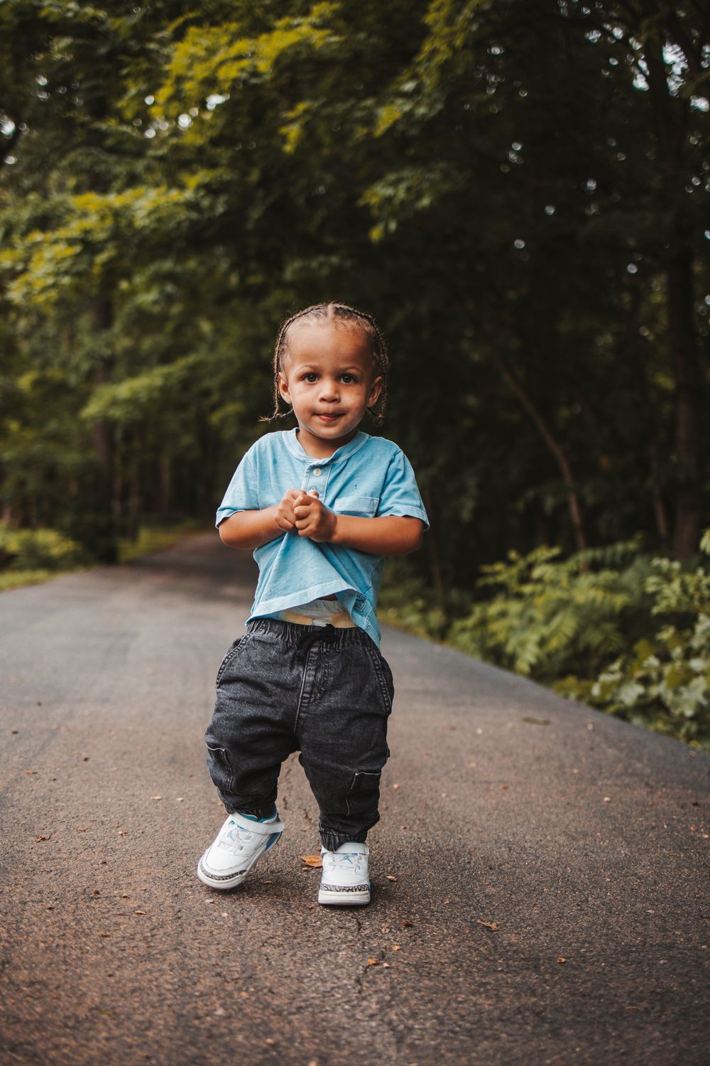 A small child in a light blue shirt and jeans stands on a tree-lined path with an excited expression.