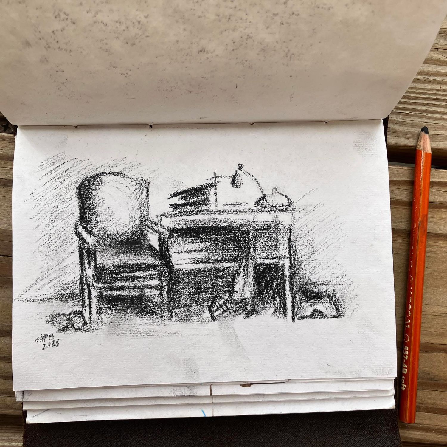 A sketchbook drawing of a cozy desk scene done in charcoal, featuring a chair, desk lamp, and scattered papers