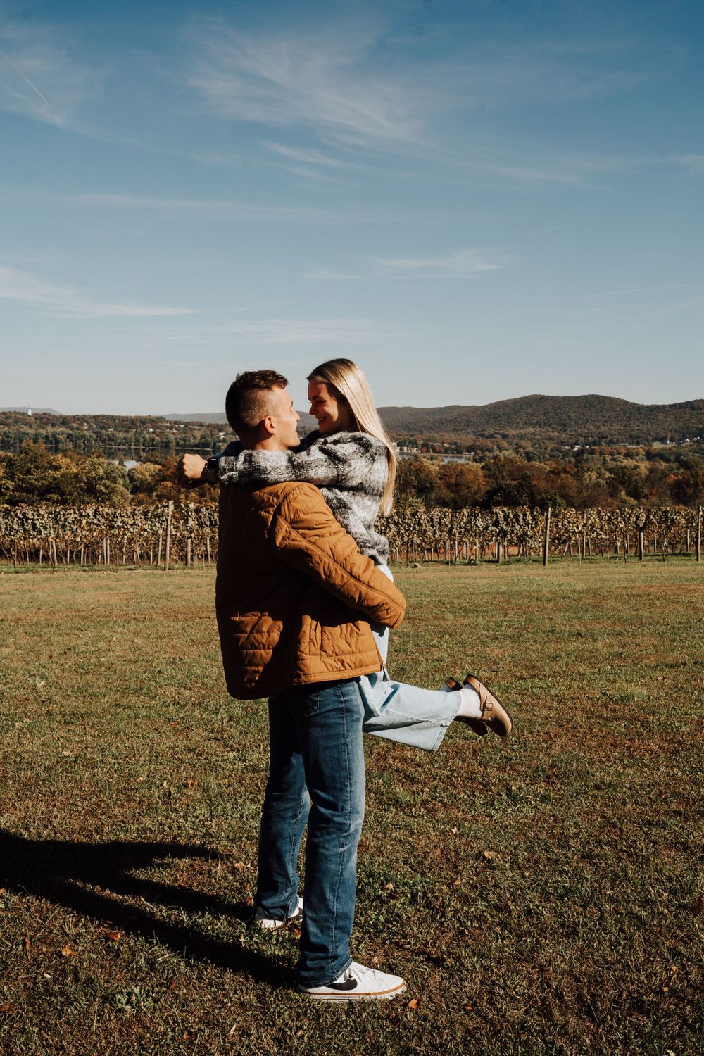 Playful couple embracing in vineyard with mountains in background.